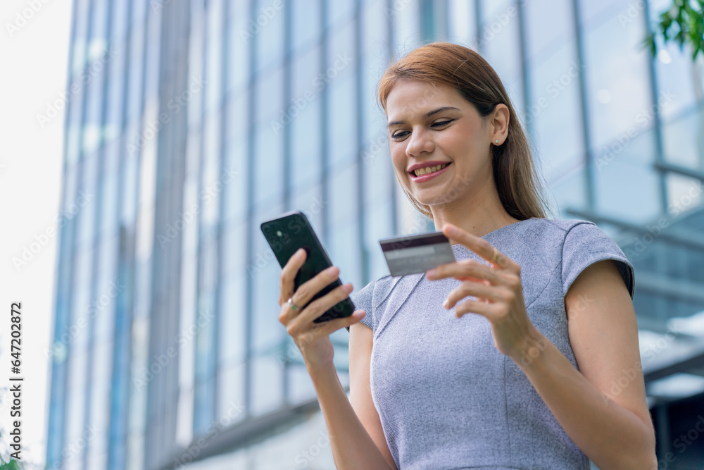 Young businesswoman doing digital banking transaction and making travel bookings using her credit card and her mobile phone in front a modern office building at a central business district