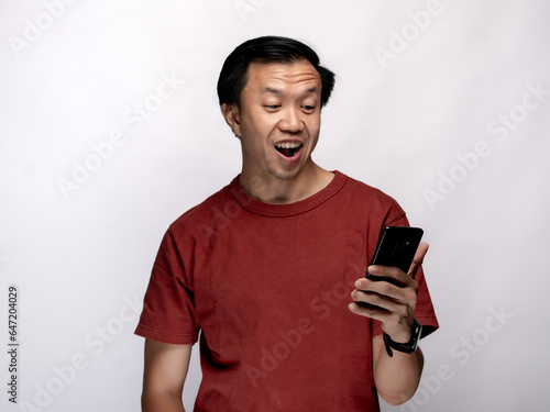 An Asian Indonesian man wearing a maroon-colored shirt is extremely delighted when looking at his smartphone, isolated against a white background.