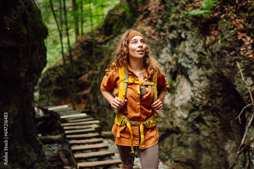 Happy woman in hiking clothes with a yellow backpack walks along a wooden hiking path in the mountains. Hiking, active lifestyle. Outdoor adventure concept. © maxbelchenko