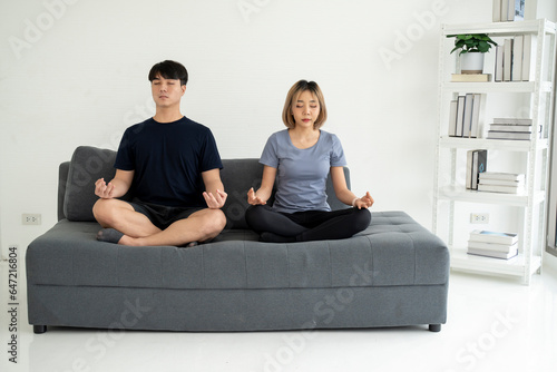 Calm young family is doing yoga together, sitting in the lotus position on the sofa.