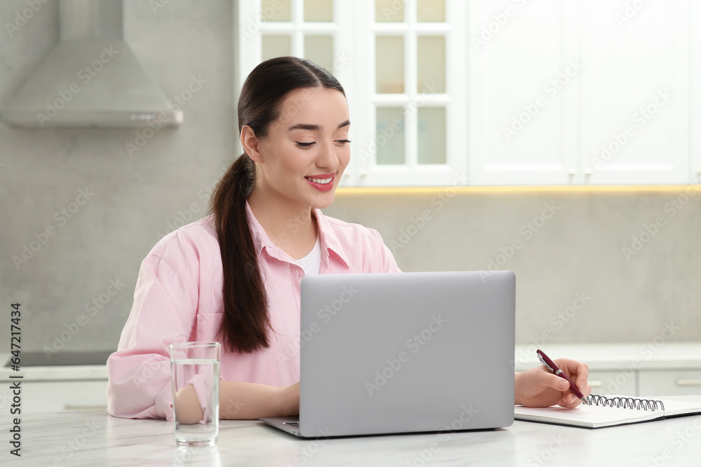 Woman writing something in notebook while using laptop at white table in kitchen Woman writing something in notebook while using laptop at white table in kitchen