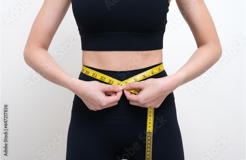 Slim Asian woman in black workout clothes Measure her waist with a yellow tape measure. She doesn't know her waist size. and checking waist size Isolated on white studio wall