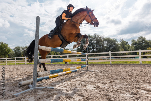 Professional equestrienne exercising hurdle jump on her beautiful horse in an outdoor riding arena with obstacles and sandy ground © 24K-Production
