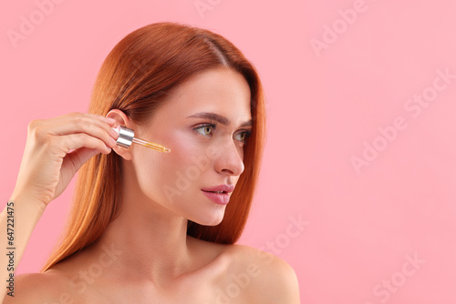Beautiful young woman applying cosmetic serum onto her face on pink background, space for text