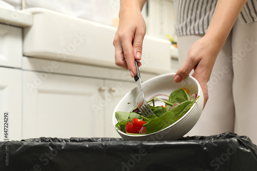 Woman throwing vegetable salad into bin indoors, closeup