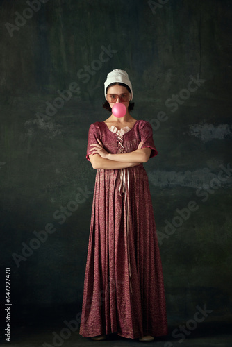 Portrait of elegant young woman in image of medieval maid, wearing historical attire and eating bubble gum on vintage green background. Concept of history, comparison of eras, beauty, art, creativity