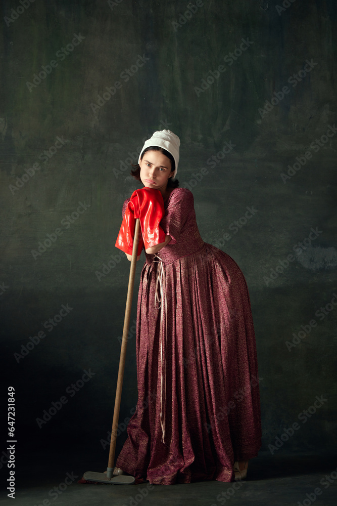 Bored and tired. Beautiful woman, medieval maid in dress standing leaning on mop in big red rubber gloves on vintage green background. Concept of history, comparison of eras, beauty, art, creativity