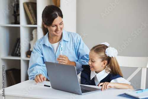 Tutor teaches little schoolgirl 7-8 years old English and gives exams on laptop, sitting at a table in a classroom during lesson. A teacher helps and explains a task to student on a computer at school