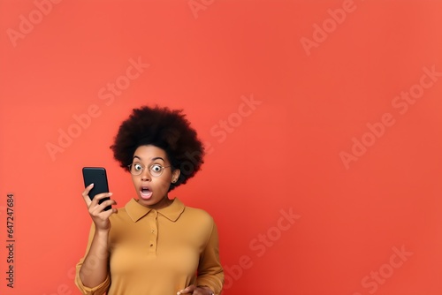 Young surprised black woman in shock holding a cellphone in front of a yellow background 