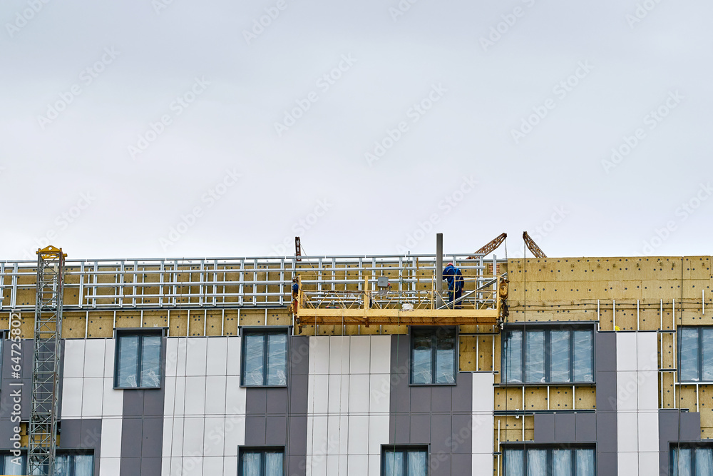 Worker on construction cradle cladding and warming wall of monolithic house. Man on suspended platform insulating wall facade of residential building, cladding system. Worker in suspended platform Worker on construction cradle cladding and warming wall of monolithic house. Man on suspended platform insulating wall facade of residential building, cladding system. Worker in suspended platform