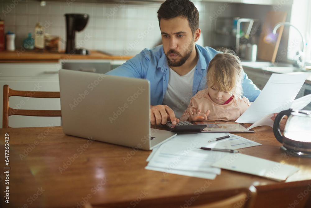 Fototapeta premium Young single father going over his bills and home finances while his daughter uses the tablet in the morning in the kitchen