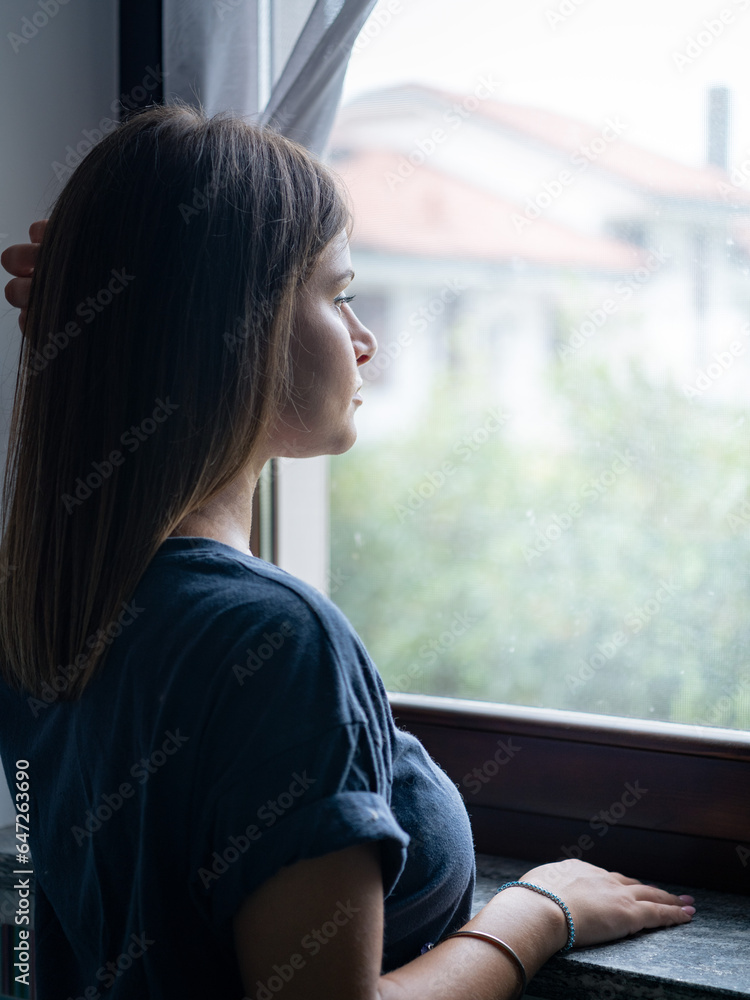 Girl looking out the window, lonely and sad with long, light brown hair. Model standing in front of the window. Girl looking out the window, lonely and sad with long, light brown hair. Model standing in front of the window.