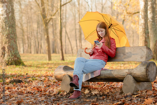 A girl in an autumn park sits on an original bench, holding an umbrella in one hand and a phone in the other.