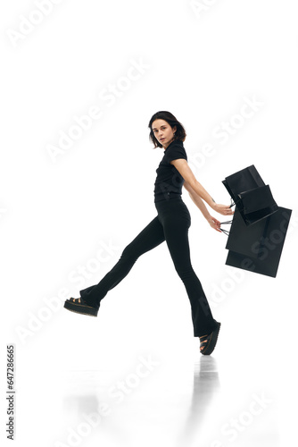 Portrait of young cheerful woman wearing fashion total black outfit holding bags in hands isolated on white studio color background. ad