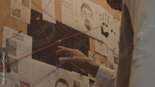 Low angle shot of Black male detective attaching old newspaper clipping to evidence corkboard while investigating criminal case in office at night