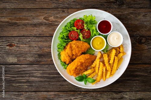 Seared breaded chicken nuggets with French fries and fresh vegetables on wooden table
