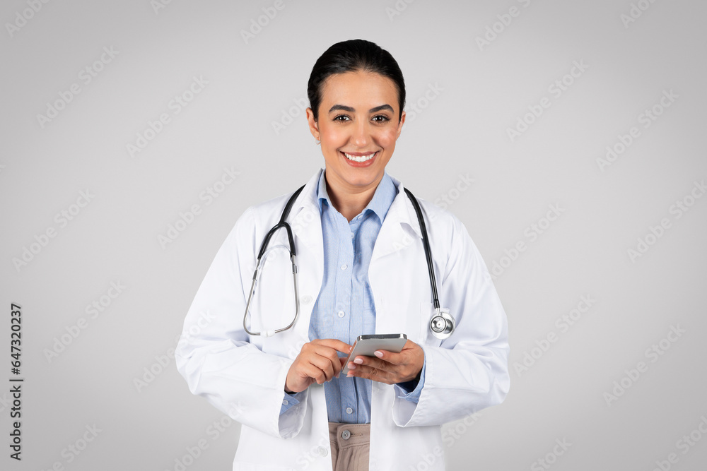Portrait of friendly latin female therapist in white coat and with stethoscope using cellphone, gray background Portrait of friendly latin female therapist in white coat and with stethoscope using cellphone, gray background
