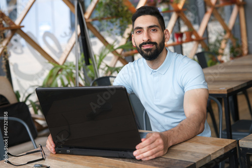 Canvas Print Portrait of Arab Muslim ethnic guy freelancer using laptop computer in glasses, young bearded man with black beard is drinking coffee sitting at table in cafe or restaurant