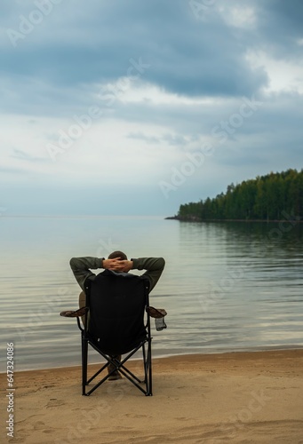 A man is sitting in a camping chair on a summer evening on the background of a forest lake.