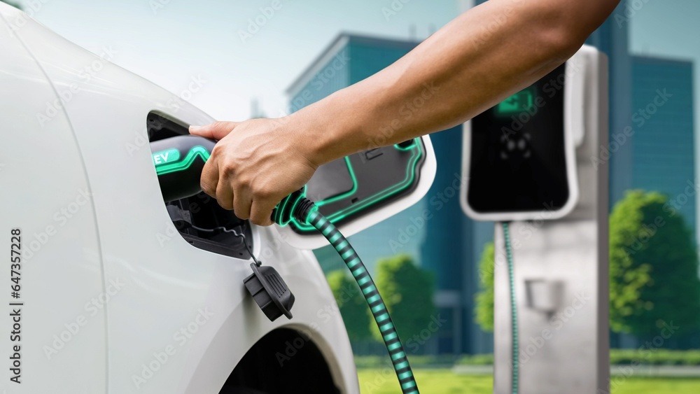 Hand insert EV charger and recharge electric car from charging station displaying futuristic battery status hologram on ESG green city park background. Smart sustainable clean energy. Peruse