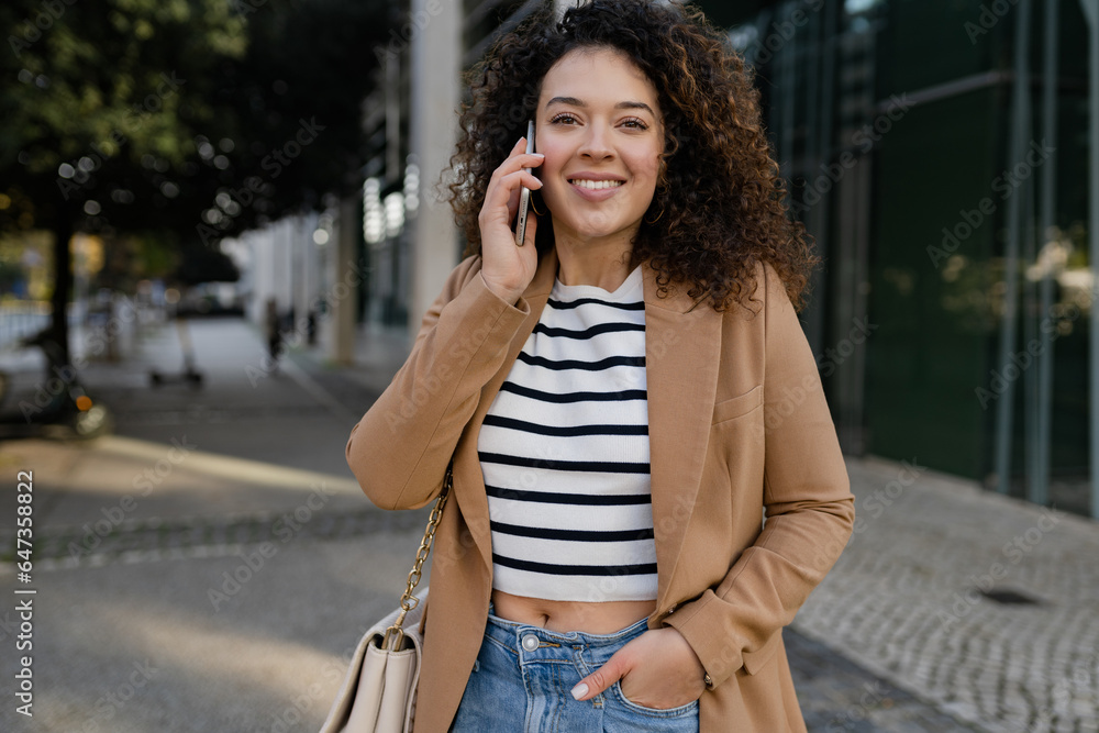 pretty curly smiling woman walking in city street in stylish jacket, talking on smartphone