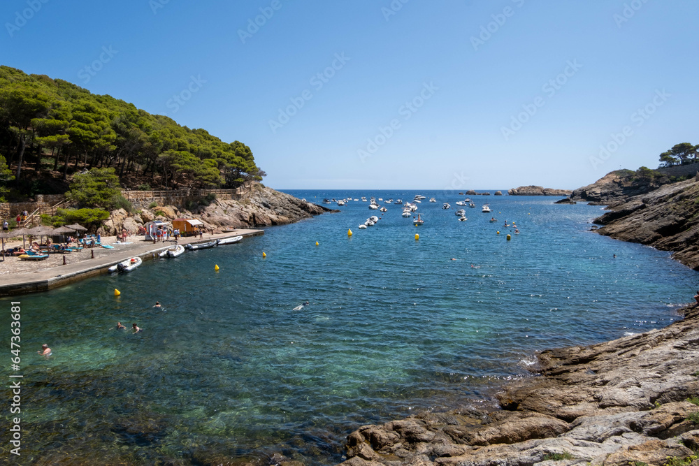 Image of a pebble beach in the Costa Brava with boats in the background.