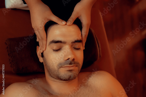 Caucasian man enjoying relaxing anti-stress head massage and pampering facial beauty skin recreation leisure in warm candle lighting ambient salon spa in luxury resort or hotel. Quiescent