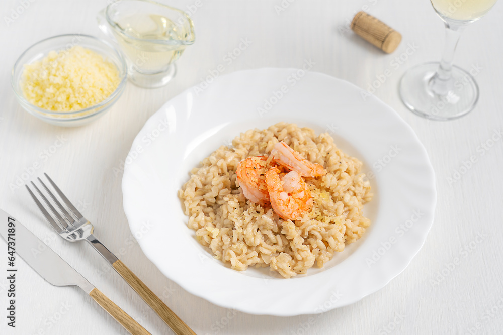 Italian homemade creamy risotto dish made of boiled rice cooked with broth decorated with shrimps or prawns served on plate on white wooden table with wine glass, tableware and grated parmesan cheese