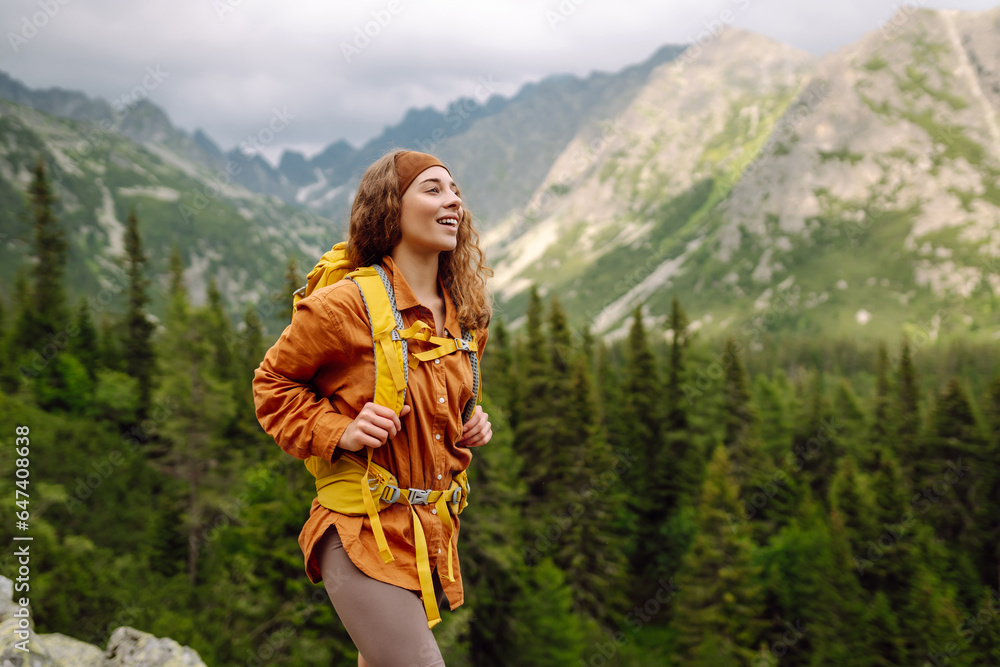 Active woman with a yellow hiking backpack traveling along hiking trails in the mountains among forests and cliffs. Traveler enjoying nature. Concept of trekking, active lifestyle. Adventures.