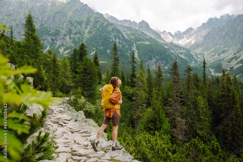 Active woman with a yellow hiking backpack traveling along hiking trails in the mountains among forests and cliffs. Traveler enjoying nature. Concept of trekking, active lifestyle. Adventures.