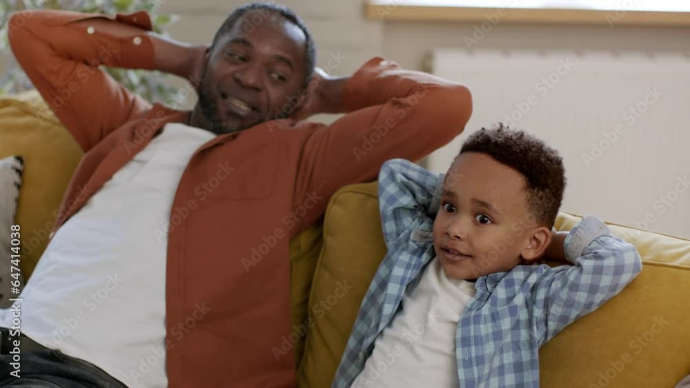 Carefree african american father and his little son resting on sofa with hands behind heads, tracking shot, slow motion Carefree african american father and his little son resting on sofa with hands behind heads, tracking shot, slow motion