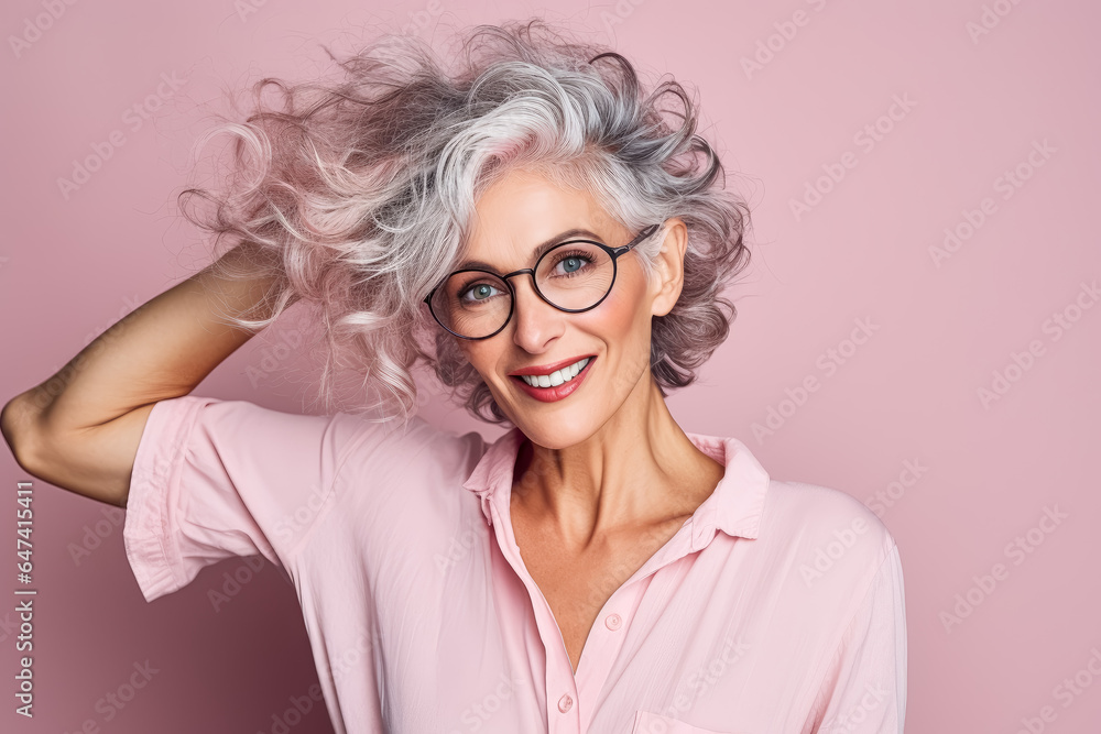 Mature Woman wearing glasses with her hand behind her hair, in front of pink background.