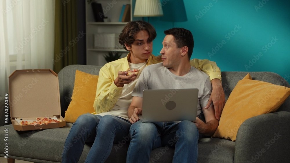 Shot of a homosexual couple at home. They are sitting on the couch, watching something on a laptop, eating pizza, hugging and talking warmly to each other. Shot of a homosexual couple at home. They are sitting on the couch, watching something on a laptop, eating pizza, hugging and talking warmly to each other.