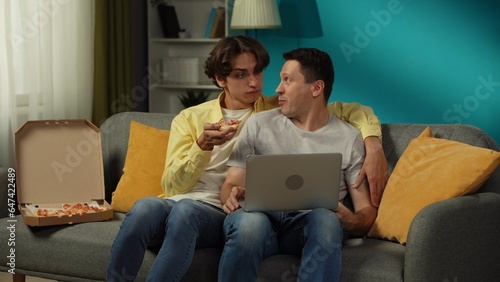 Shot of a homosexual couple at home. They are sitting on the couch, watching something on a laptop, eating pizza, hugging and talking warmly to each other.