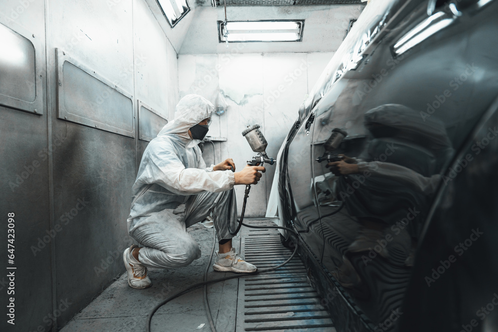 Automotive service worker in full protective gear expertly apply color paint in to car's bodywork with spray gun or respirator painting in chamber workshop. Car paint service for scratch refinish.Oxus