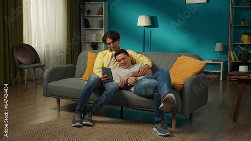 Full-size shot of a homosexual couple at home. They are laying on the couch, watching photos or videos on a tablet, hugging, smiling and expressing love to each other.