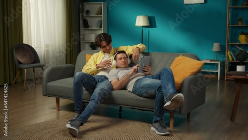 Full-size shot of a homosexual couple at home. They are laying on the couch, watching photos or videos on a tablet. One is sharing their headphones.