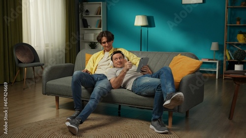 Full-size shot of a homosexual couple at home. They are laying on the couch, watching photos or videos on a tablet, hugging, smiling. One is listening to music.