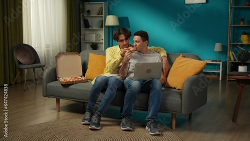 Full-size shot of a homosexual couple at home. They are sitting on the couch, watching something on a laptop, eating pizza, hugging and talking warmly to each other.