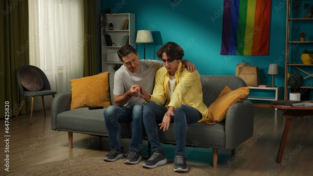 Full-size shot of a homosexual, pansexual, bisexual male couple sitting on the couch, hugging, holding hands. One is comforting their partner. Full-size shot of a homosexual, pansexual, bisexual male couple sitting on the couch, hugging, holding hands. One is comforting their partner.