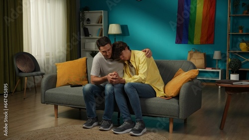 Shot of a homosexual male couple standing in front of each other, looking at each other and holding their hands tenderly. Rainbow flag on the background.