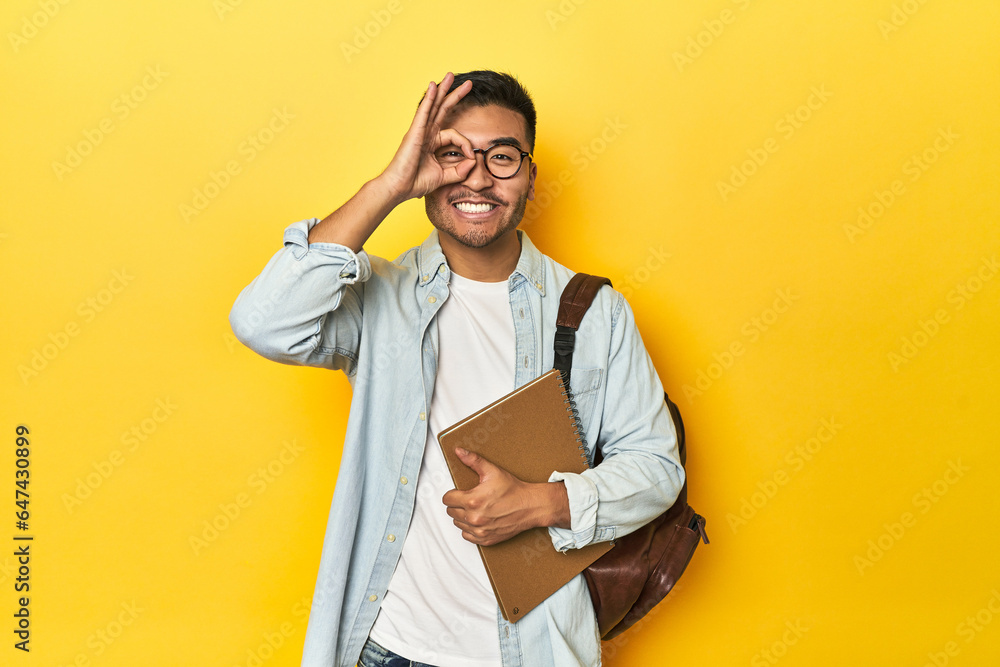 Asian student with backpack and notebook, yellow studio backdrop excited keeping ok gesture on eye. - obrazy, fototapety, plakaty 