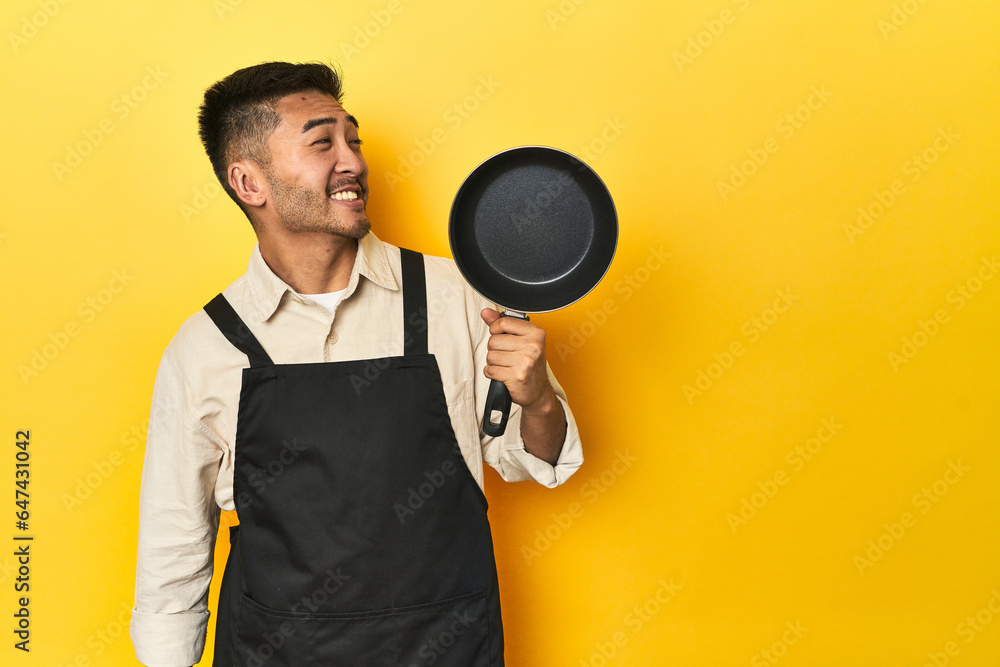 Asian chef holding a pan, yellow studio backdrop looks aside smiling, cheerful and pleasant. Asian chef holding a pan, yellow studio backdrop looks aside smiling, cheerful and pleasant.
