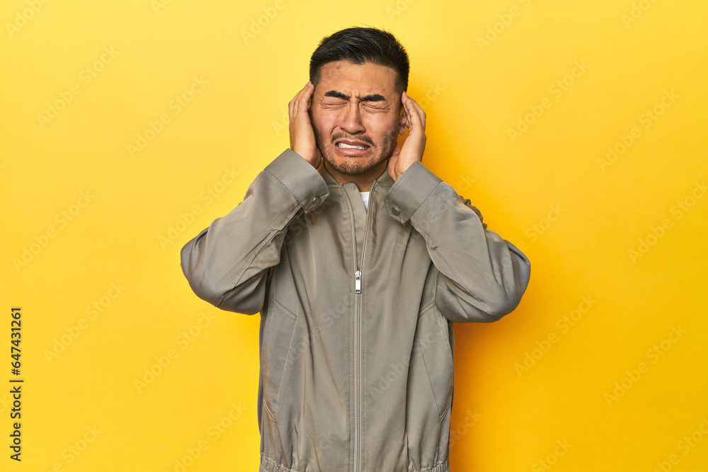 Stylish Asian man in gray jacket on yellow studio covering ears with hands. Stylish Asian man in gray jacket on yellow studio covering ears with hands.
