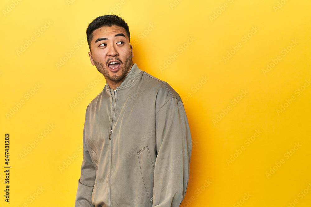Stylish Asian man in gray jacket on yellow studio looks aside smiling, cheerful and pleasant.