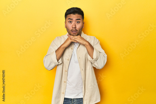 Casual Asian man with open shirt, white tee on yellow studio praying for luck, amazed and opening mouth looking to front. © Asier