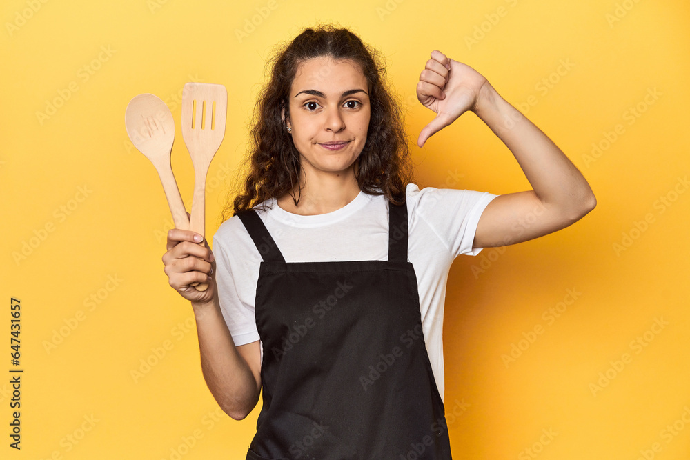 Woman with apron, wooden cooking utensils, yellow, showing a dislike gesture, thumbs down. Disagreement concept. Woman with apron, wooden cooking utensils, yellow, showing a dislike gesture, thumbs down. Disagreement concept.