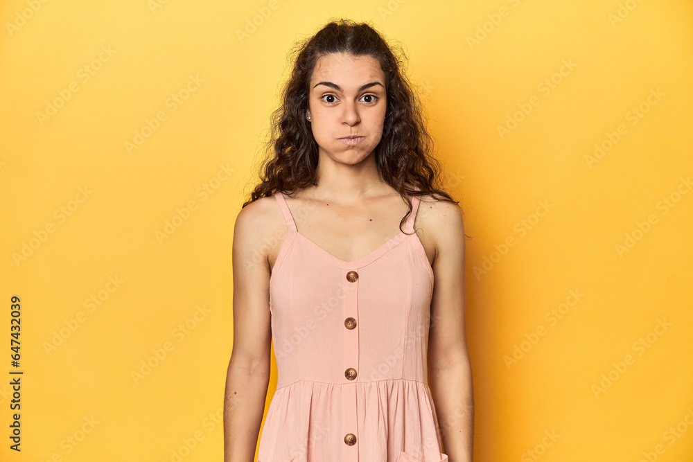 Young Caucasian woman, yellow studio background, blows cheeks, has tired expression. Facial expression concept. - obrazy, fototapety, plakaty 