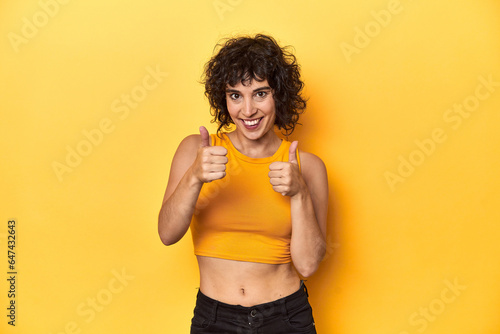 Curly-haired Caucasian woman in yellow top raising both thumbs up, smiling and confident.