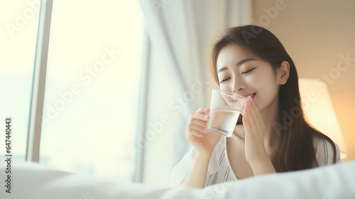 Asian young beautiful woman sitting on bed holding water of glass and drinking in the morning in bedroom at home after wake up in the morning